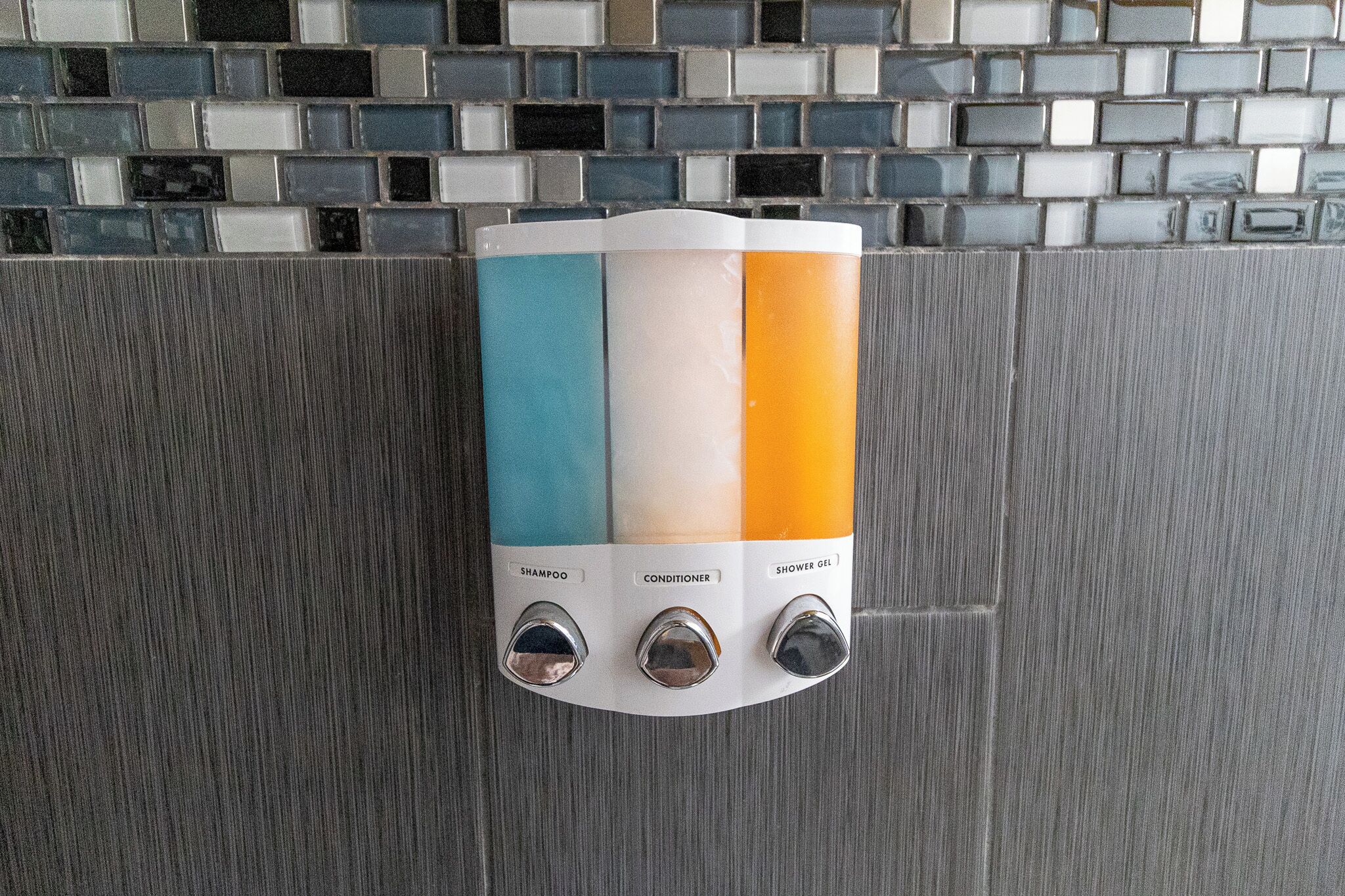 Eine wandmontierte Spenderstation mit drei Behältern, beschriftet für Shampoo, Conditioner und Duschgel.