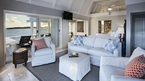 Un salon moderne avec un grand canapé blanc, un petit pouf et une table d’appoint en osier. Une grande fenêtre offre une vue sur la mer.