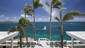 Une vue sur la plage, avec des palmiers, un voilier et un espace de détente blanc avec des chaises.