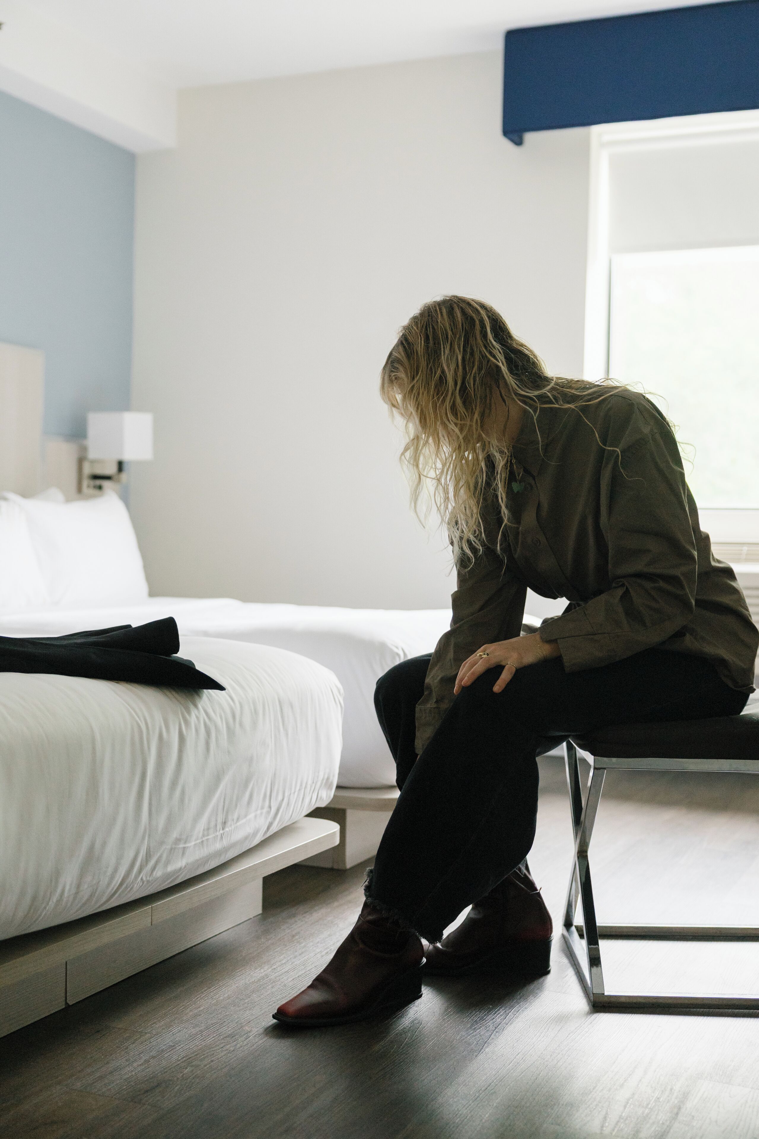 A person sitting on a chair in a room with a bed, a nightstand, and a lamp.