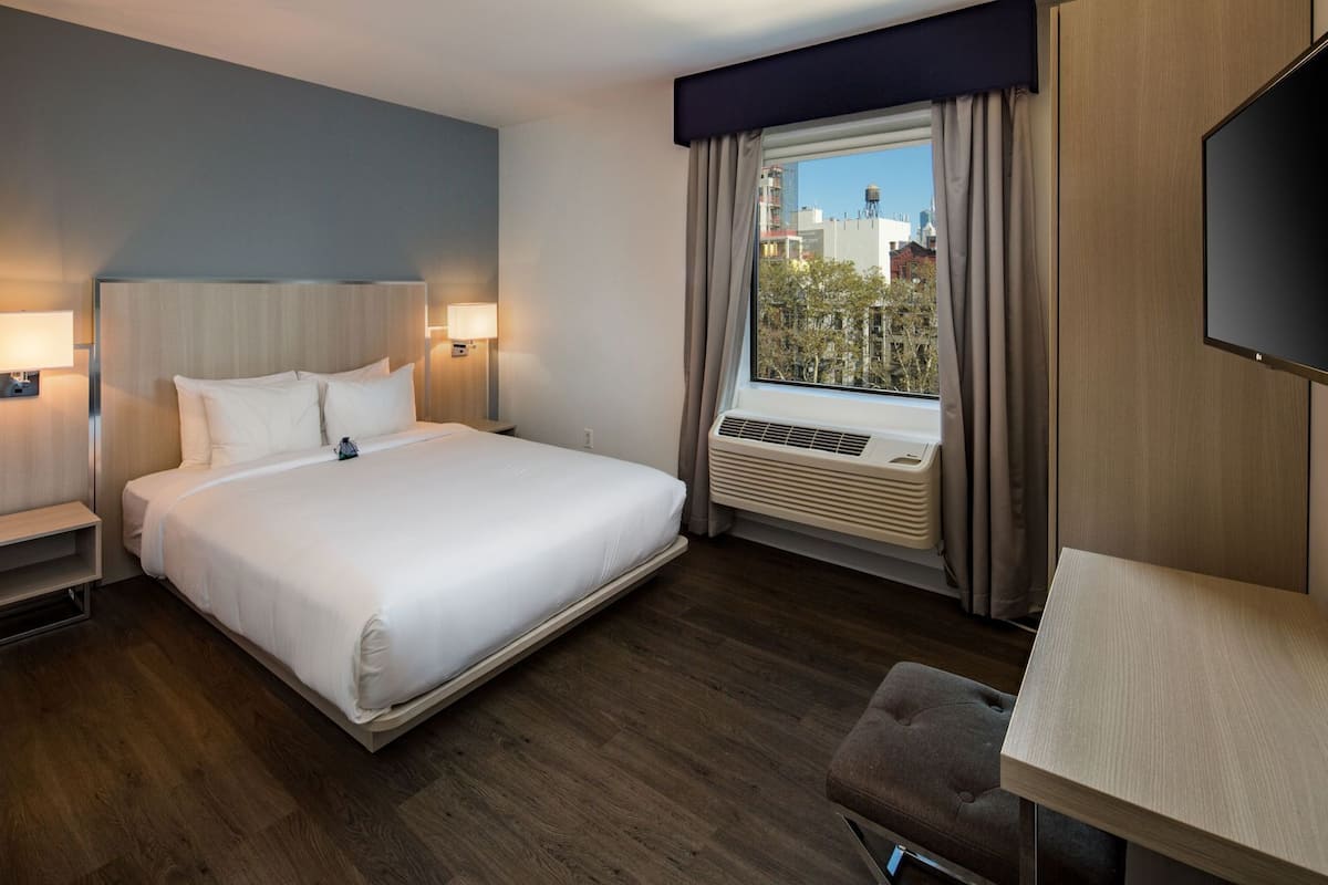Habitación de hotel con cama, mesitas de noche, televisión, ventana con cortinas y aire acondicionado.