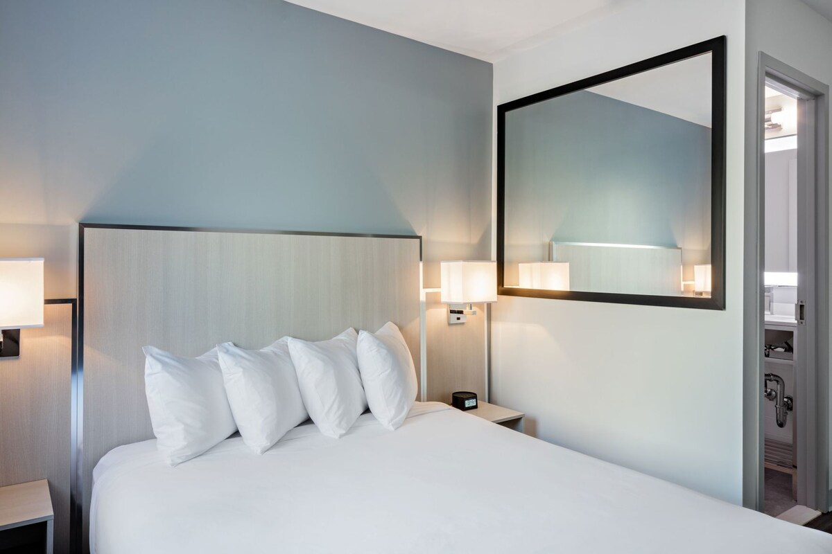 A hotel room with a large bed, bedside lamps, a nightstand, a mirror, and a door leading to a bathroom.