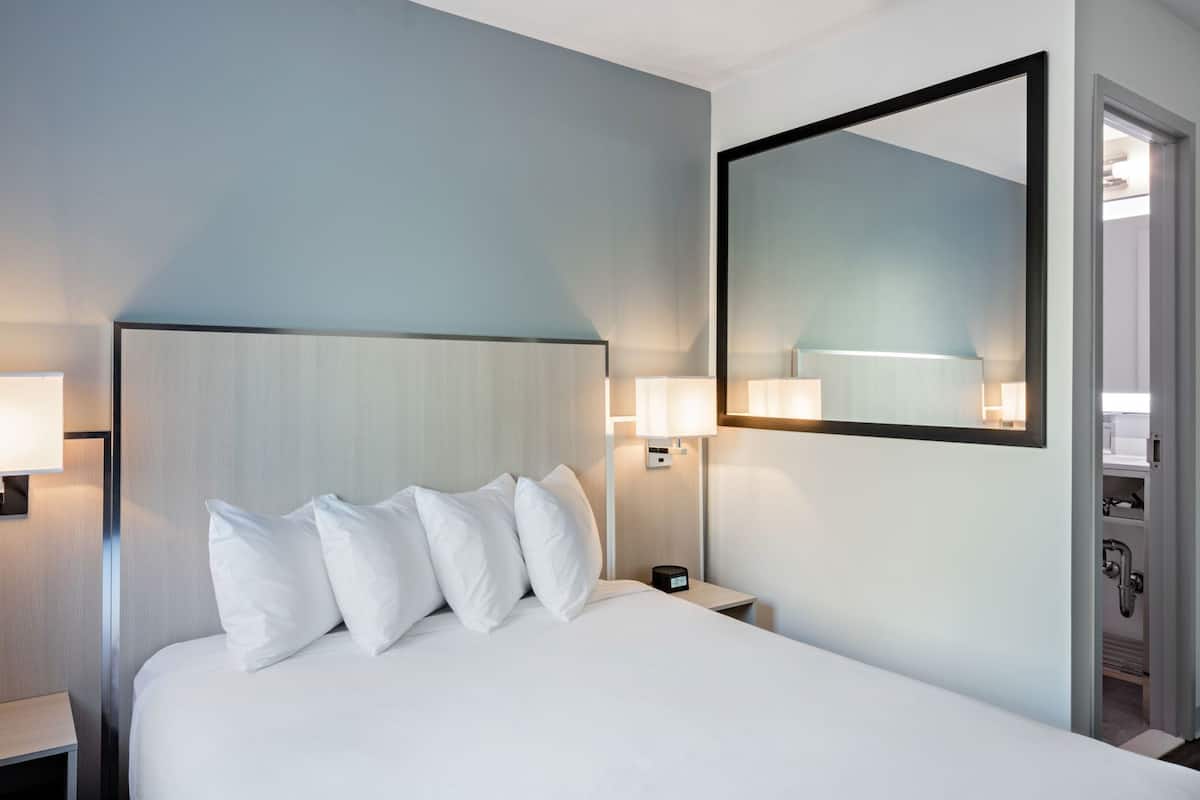 Habitación de hotel con una cama grande, lámparas de noche, una mesita de noche, un espejo y una puerta que da al baño.