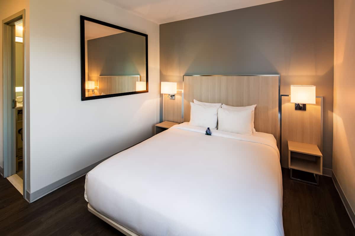 Una habitación de hotel con una cama grande, dos mesitas de noche con lámparas, un espejo y un cabecero de madera.