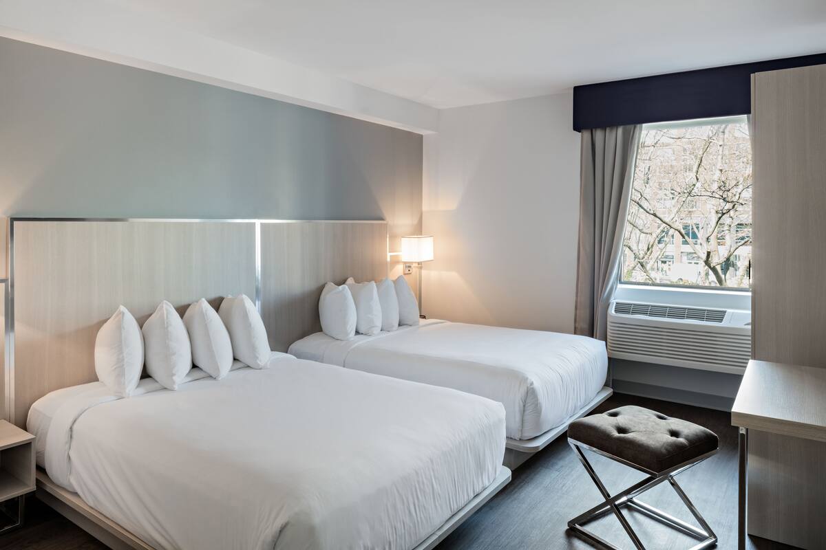 Habitación de hotel con dos camas, un escritorio, una lámpara y una ventana con vista a árboles.