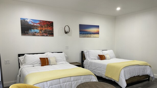 Habitación de hotel con dos camas, un sillón amarillo, una mesita de madera y dos cuadros en la pared.