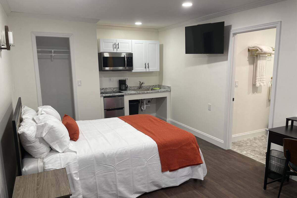 A compact hotel room with a bed, a small desk, a kitchenette, and a TV.