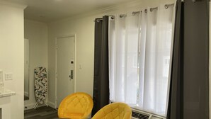 Una habitación con dos sillones amarillos, una pequeña mesa de madera, una puerta blanca y una ventana con cortinas grises.