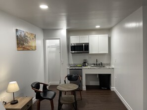 A compact kitchen with white cabinets, a microwave, and a sink. There is a small dining area with two chairs and a table. A painting of a beach scene hangs on the wall.