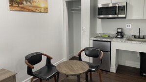 Una cocina compacta con armarios blancos, un microondas de acero inoxidable y una cafetera. Hay dos sillas negras y una pequeña mesa de madera. Un cuadro con una escena de playa cuelga en la pared.