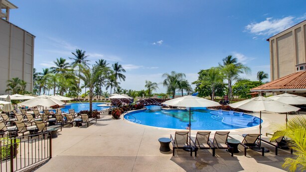 A resort pool area with lounge chairs, umbrellas, and palm trees. - The Palms 101 (Jacó)