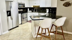 Una cocina moderna con armarios blancos, electrodomésticos de acero inoxidable, una encimera negra y dos taburetes blancos.