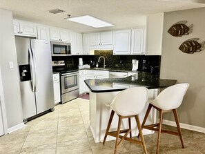 A modern kitchen with white cabinetry, stainless steel appliances, a black countertop, and two white bar stools. - Unbelievable Views From This Beautiful 3 Bedroom Condo on the 7th Floor! (Orange Beach)
