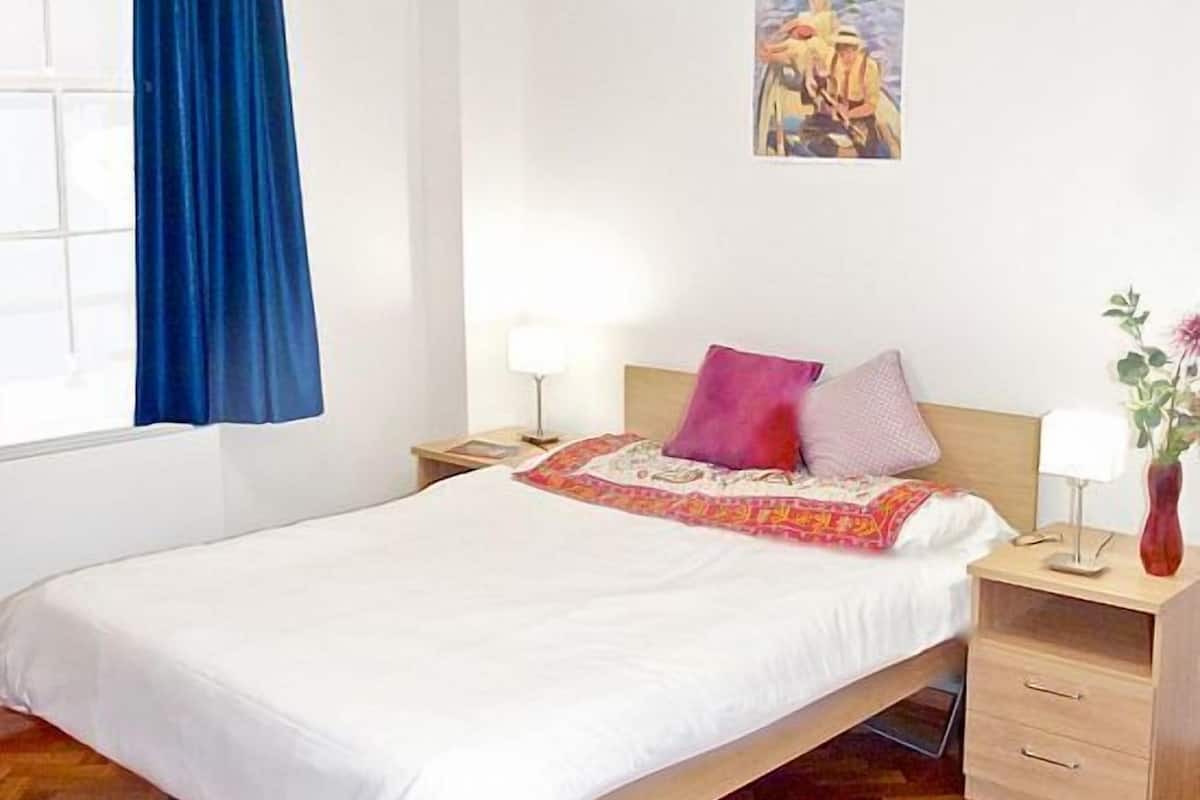 Un dormitorio con una cama, dos mesitas de noche, una lámpara auxiliar, una ventana con cortinas azules y un cuadro en la pared.