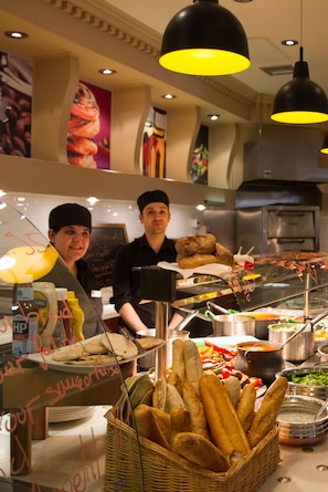 Ein Buffetbereich mit verschiedenen Speisen, darunter Brot und Soßen, sowie zwei Mitarbeiter hinter der Theke.