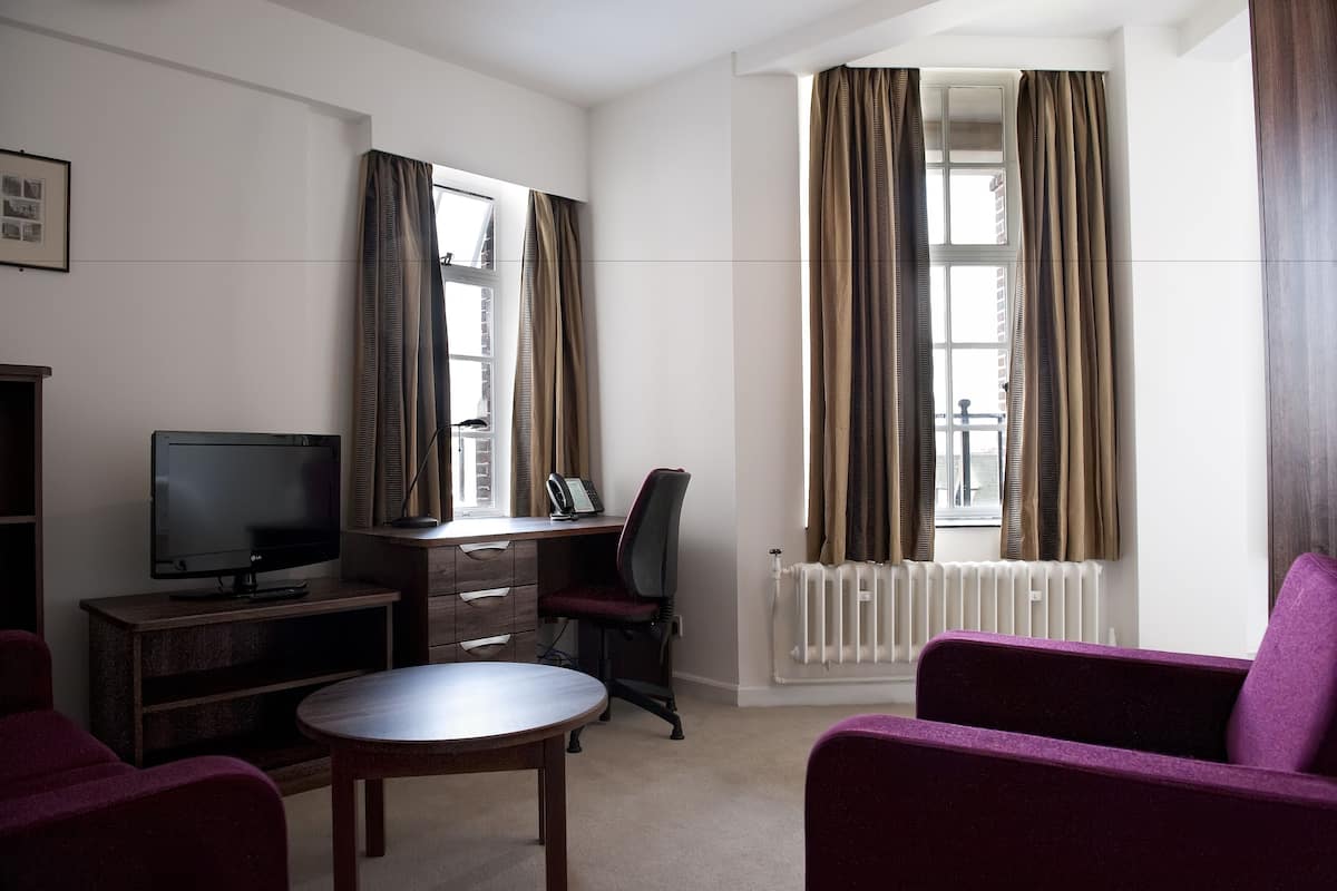 Habitación de hotel con televisor, escritorio, silla, sofá púrpura y ventanas amplias con cortinas.