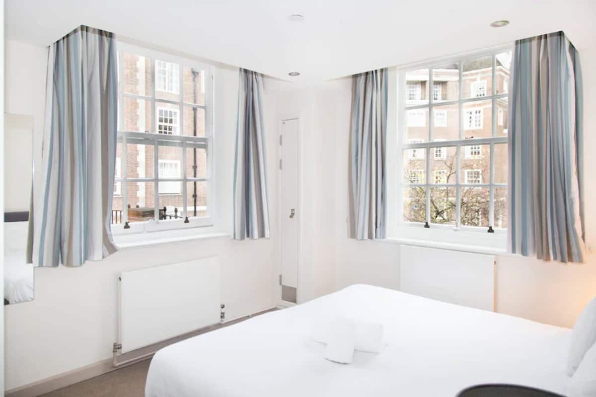 A hotel room with a large bed, two windows with curtains, a radiator, and a view of buildings outside.