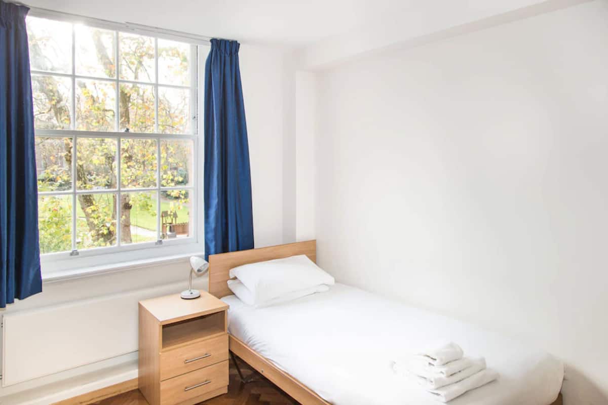 Un dormitorio pequeño y ordenado con una cama individual, una mesita de noche con lámpara y una ventana con cortinas.