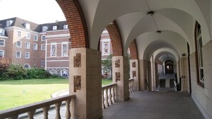Ein überdachter Gang mit Bögen, ein Backsteingebäude im Hintergrund und eine Rasenfläche.