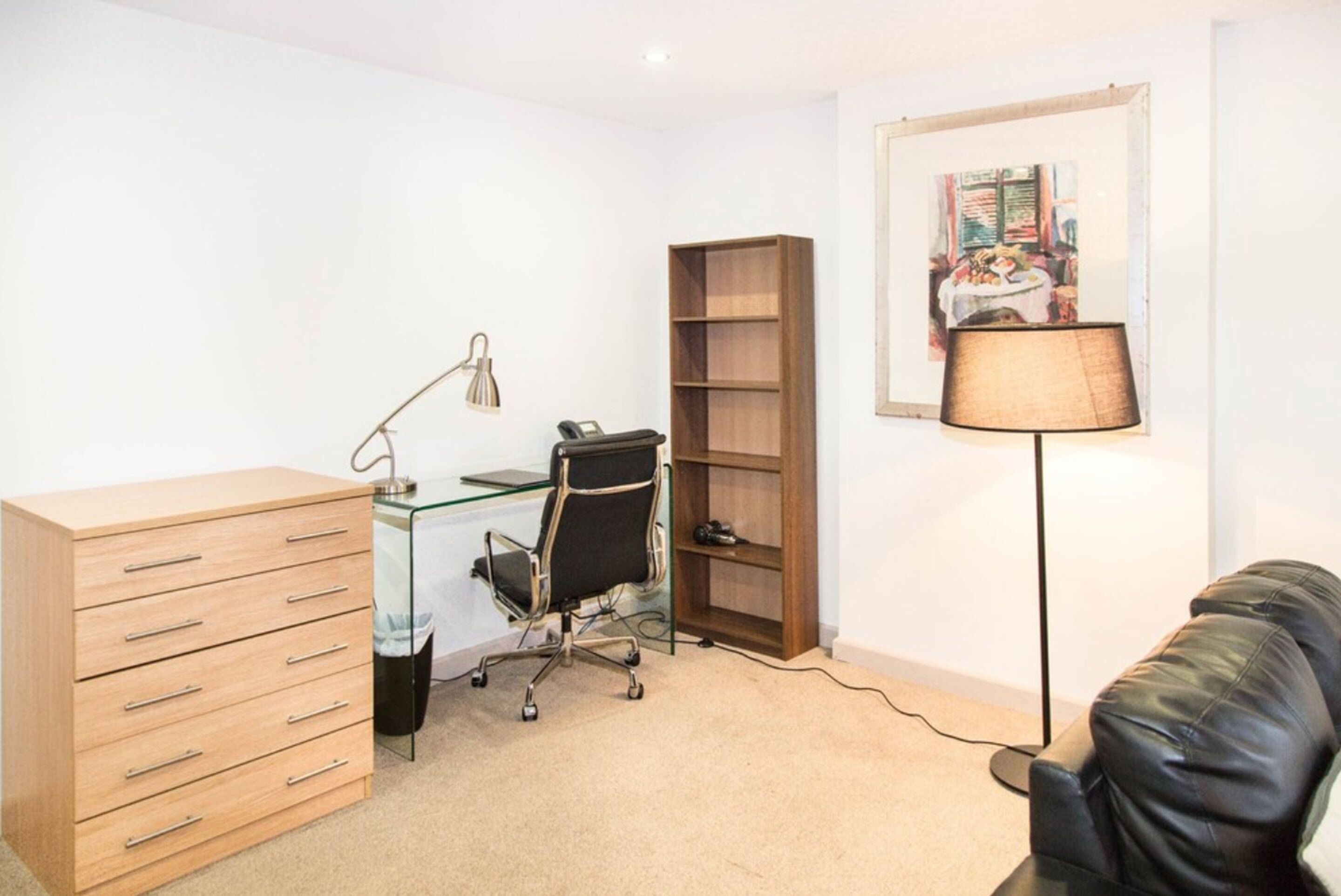 Une pièce comprenant une commode en bois, un bureau en verre avec un fauteuil de bureau noir, une bibliothèque et un lampadaire.