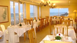Un restaurant avec des tables dressées pour le dîner, de grandes fenêtres donnant sur la mer et un lustre.