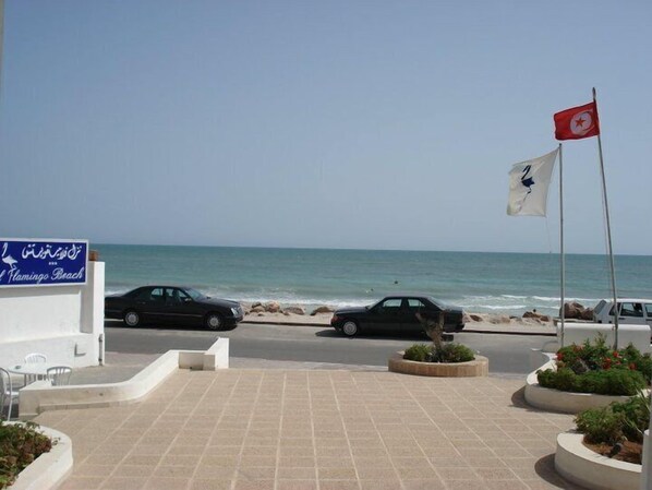 Zona frente al mar con un cartel que indica Playa Flamingo, un estacionamiento con coches y un asta con bandera.