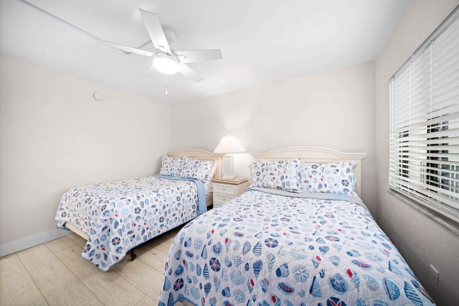 Une chambre avec deux lits, un ventilateur de plafond, une lampe de chevet et une fenêtre avec des stores.