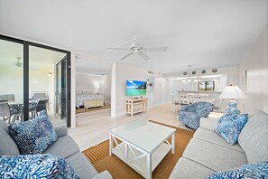 A modern living room with a glass partition, a dining area, a kitchen, and a bedroom with a TV.