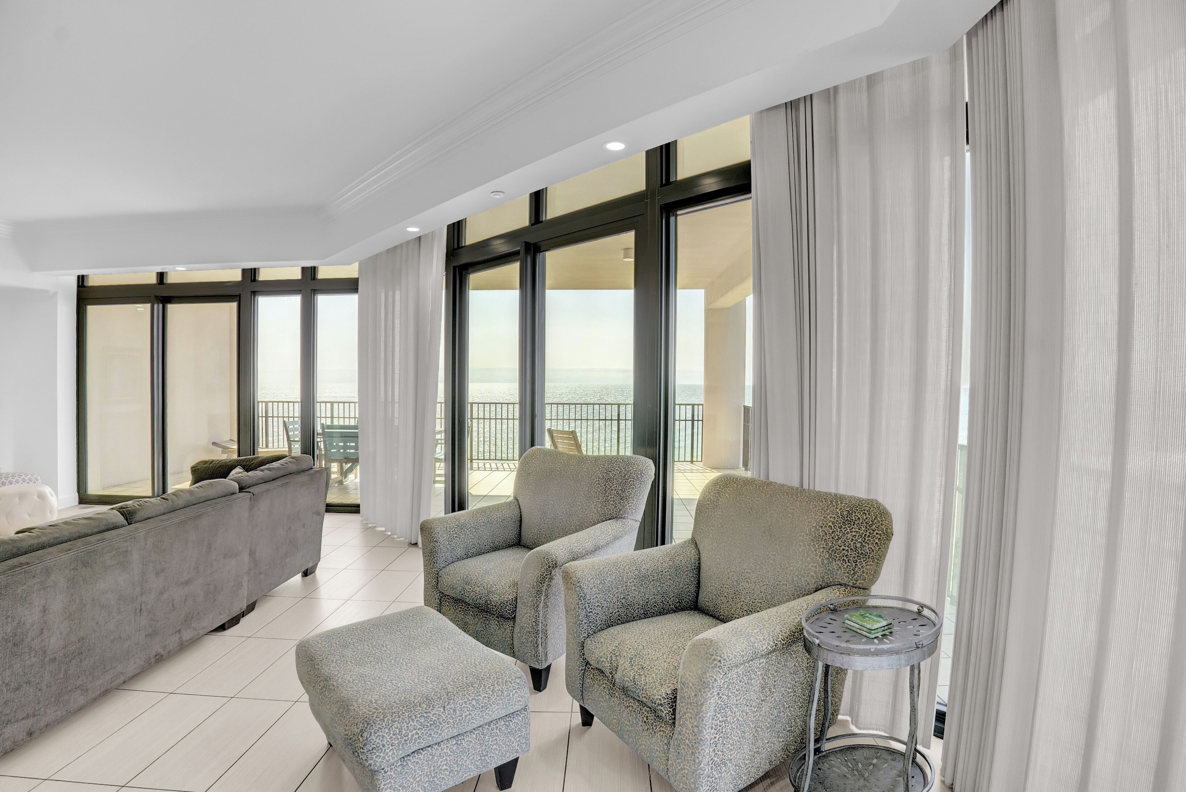 Amplia sala de estar con un sofá gris, dos sillones y una mesita auxiliar. Grandes ventanales ofrecen vistas al mar.