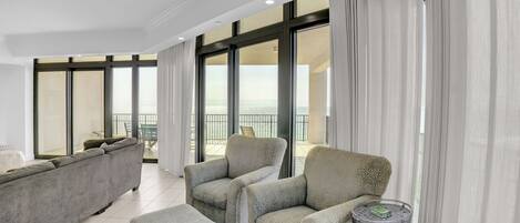 Amplia sala de estar con un sofá gris, dos sillones y una mesita auxiliar. Grandes ventanales ofrecen vistas al mar.