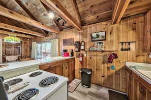 A wooden cabin interior with a kitchen area, including a stove, cabinets, and a hanging rack with utensils.