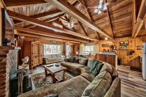 A rustic cabin interior with a large wooden ceiling, a cozy living area with a sofa, a dining table, and a kitchen area with wooden cabinets and a refrigerator.