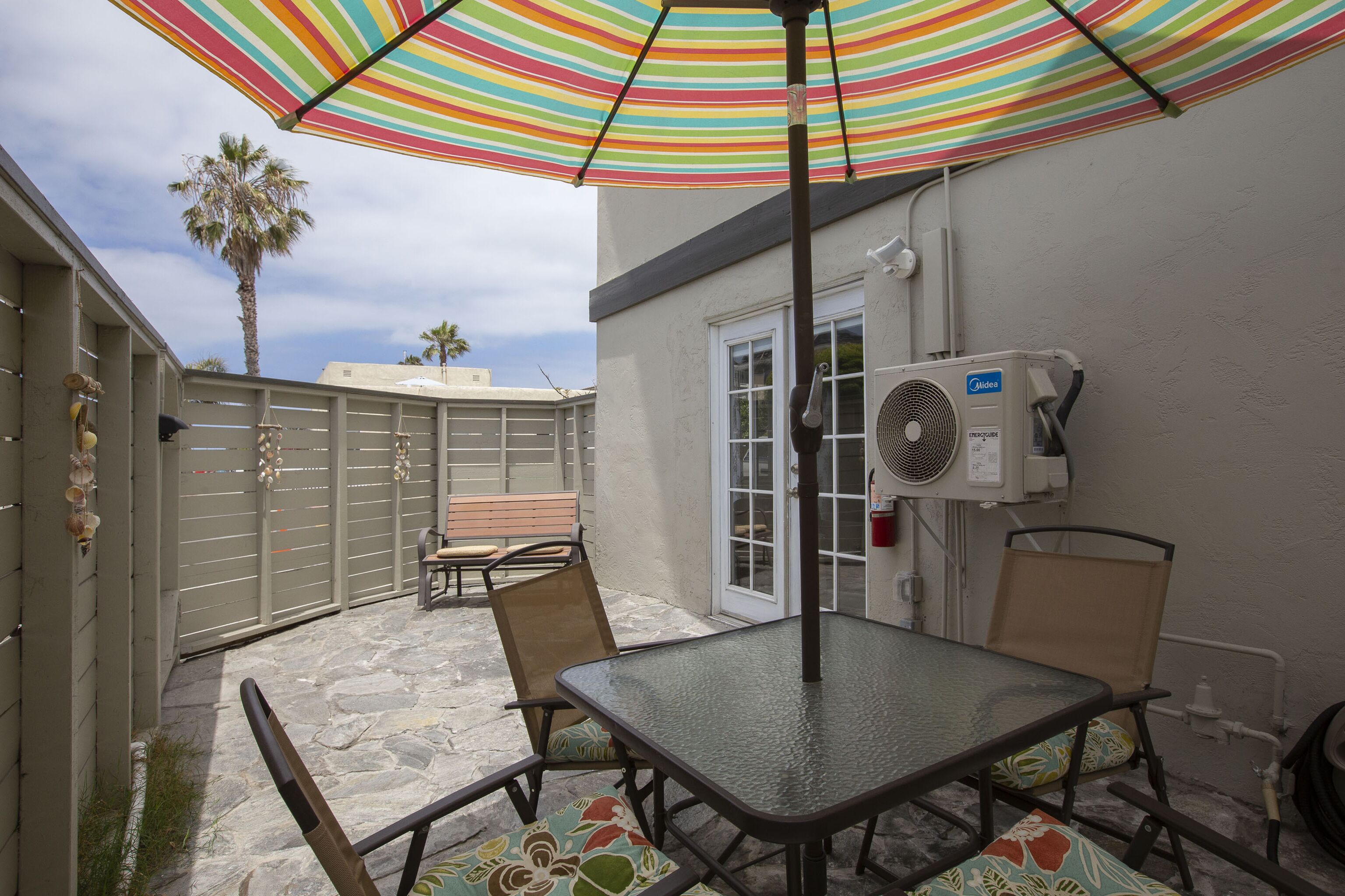 A patio area with a table and chairs, a striped umbrella, a wall-mounted air conditioning unit, and a gate with decorative items.