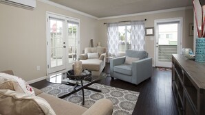 Un salon avec un canapé beige, un fauteuil bleu clair, une table basse en verre et un tapis à motifs. Il y a deux portes donnant sur l’extérieur, l’une avec un panneau de verre et l’autre avec une porte blanche.