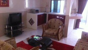 A living room with a sofa, a coffee table, a TV, a patterned armchair, and a small kitchen area with a microwave. - Residence La Suite (St. Louis)