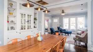 A spacious dining area with a wooden table and orange chairs, a kitchen with white cabinets, and a living room with a blue sofa and a ceiling fan. - Garrett House | Hill Country retreat Kerrville - pool - hot tub - outdoor shower (Kerrville)
