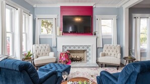 A living room with a fireplace, two armchairs, a blue sofa, and a TV mounted above the fireplace. - Garrett House | Hill Country retreat Kerrville - pool - hot tub - outdoor shower (Kerrville)