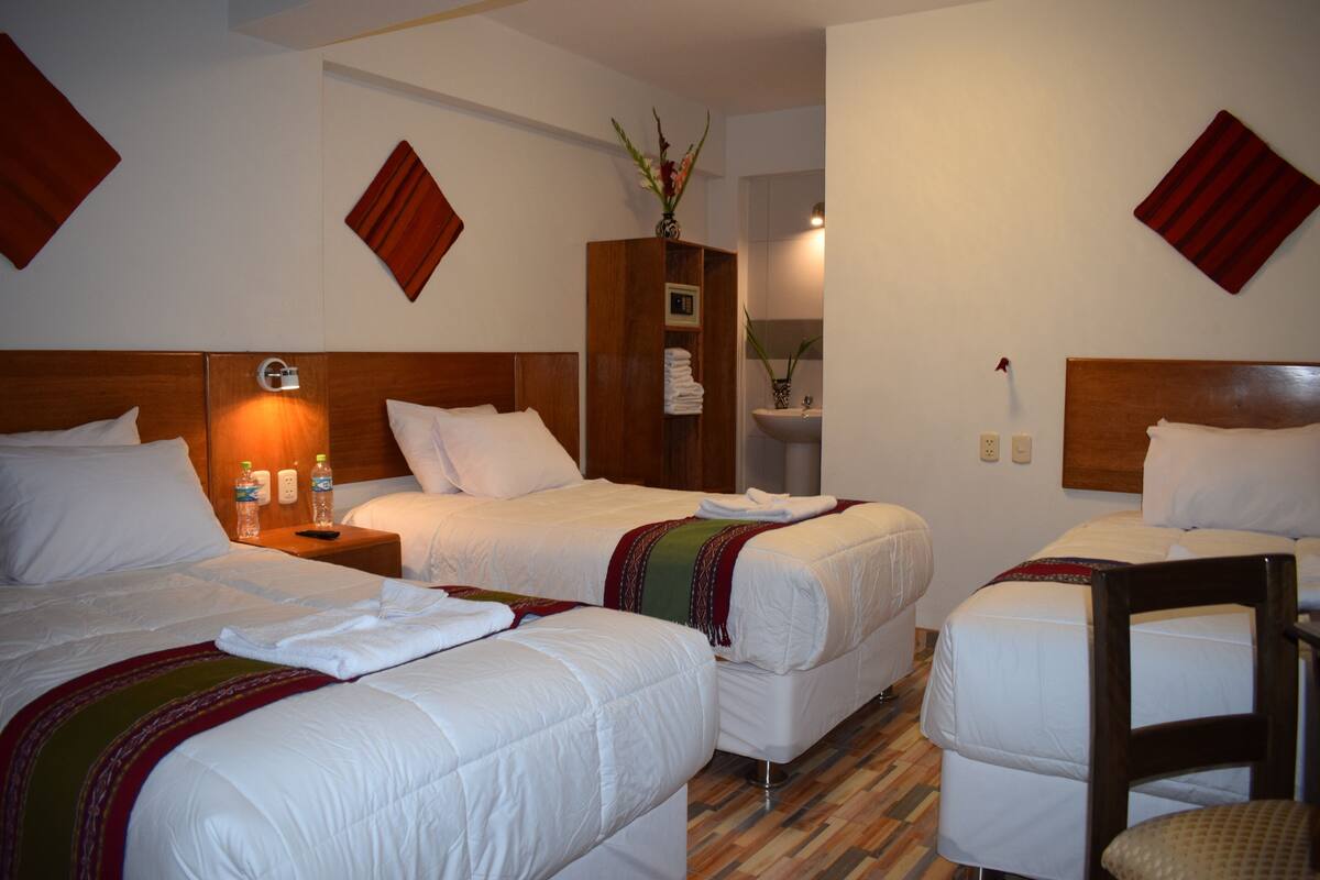 A hotel room with three single beds, a wooden headboard, a nightstand with a lamp, and a small wooden cabinet with a vase.