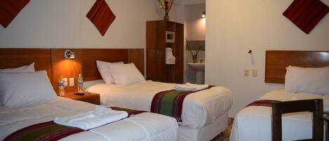 Habitación de hotel con tres camas individuales, un cabecero de madera, una mesita de noche con lámpara y un pequeño armario de madera con un jarrón.