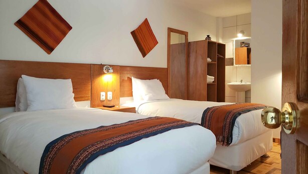 Habitación de hotel con dos camas, cabeceros de madera, una mesita de noche con lámpara, un armario y un baño visible a través de una puerta abierta.