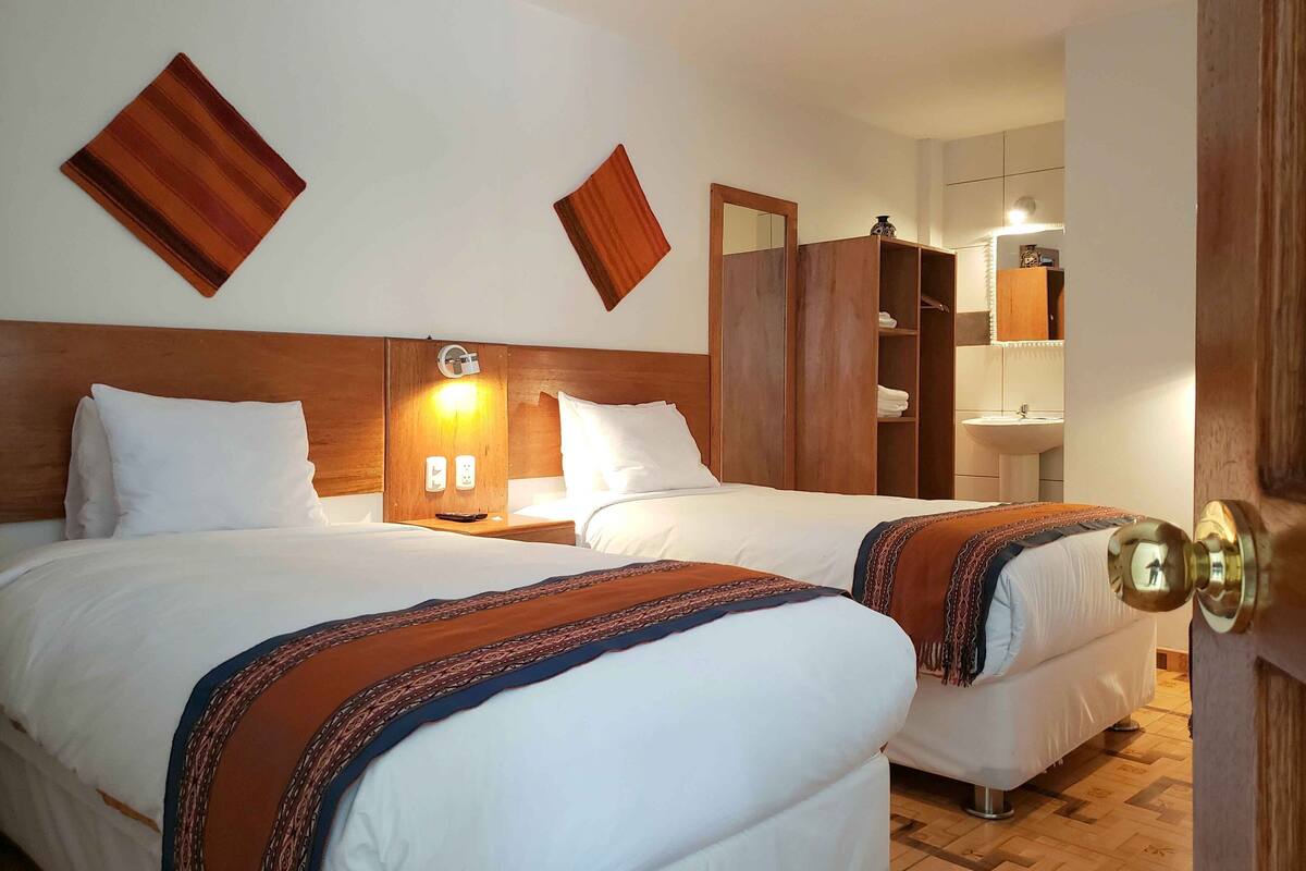 A hotel room with two beds, wooden headboards, a nightstand with a lamp, a wardrobe, and a bathroom visible through an open door.