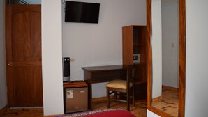 Un dormitorio con un armario de madera, un escritorio con silla, un televisor montado en la pared y un refrigerador pequeño.