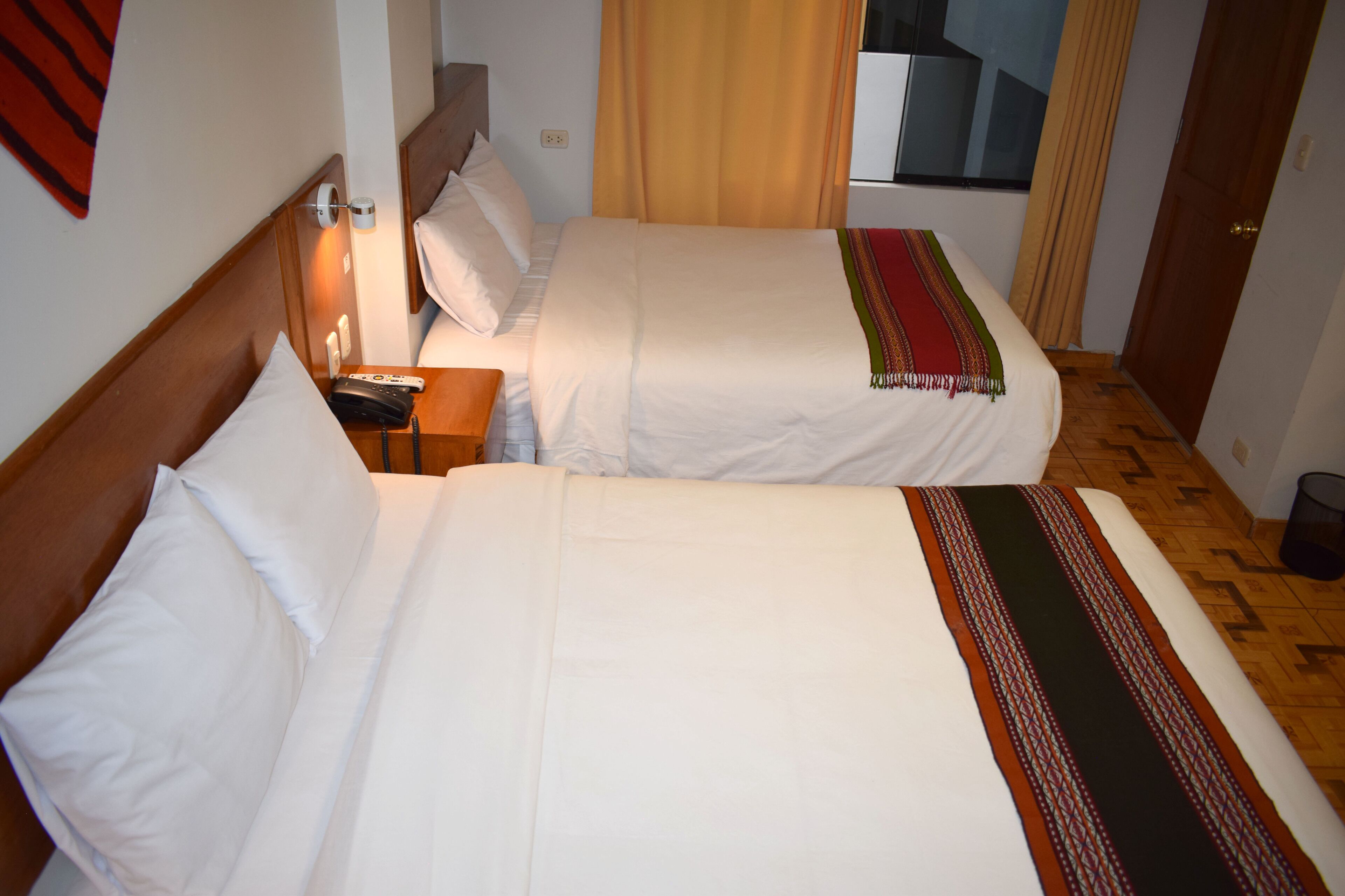 Habitación de hotel con dos camas individuales, cabecera de madera, mesita de noche con lámpara y ventana con cortinas.