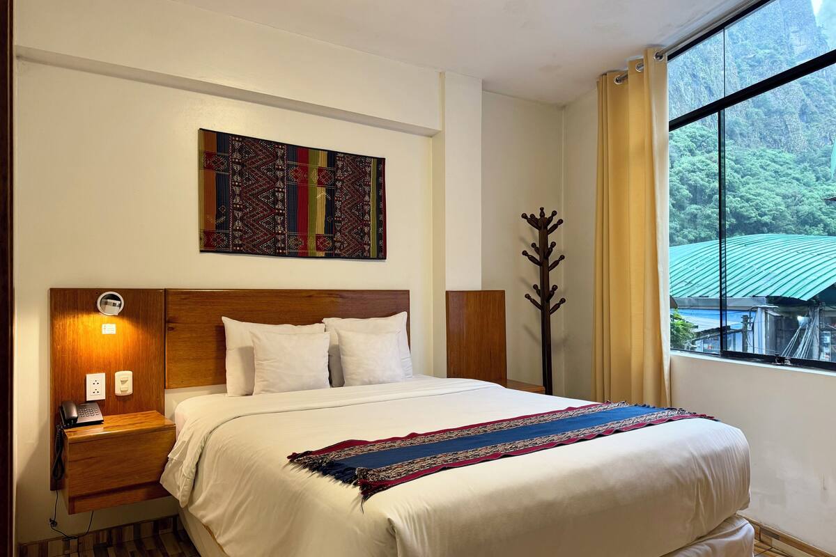A hotel room with a wooden headboard, a bed with white linens, a colorful artwork above the bed, a nightstand with a phone and a lamp, and a window with a view of greenery and a building.