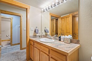 Une salle de bain avec un meuble-lavabo en bois, un évier et un miroir. Des serviettes sont accrochées à des crochets, et un flacon de savon liquide est posé sur le comptoir.