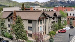 A multi-story building with a parking lot in front, surrounded by trees and mountains in the background.