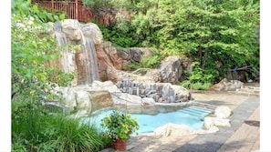 Una piscina natural de roca con cascada, rodeada de exuberante vegetación y una terraza de madera.