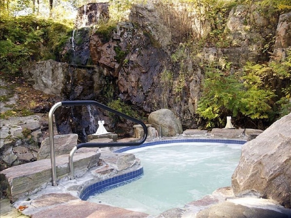 Un manantial caliente rodeado de rocas helechos, con una escalera metálica que baja al agua.
