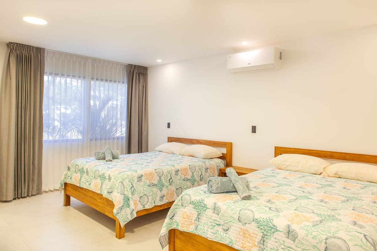A hotel room with two beds, a window with curtains, and an air conditioning unit.