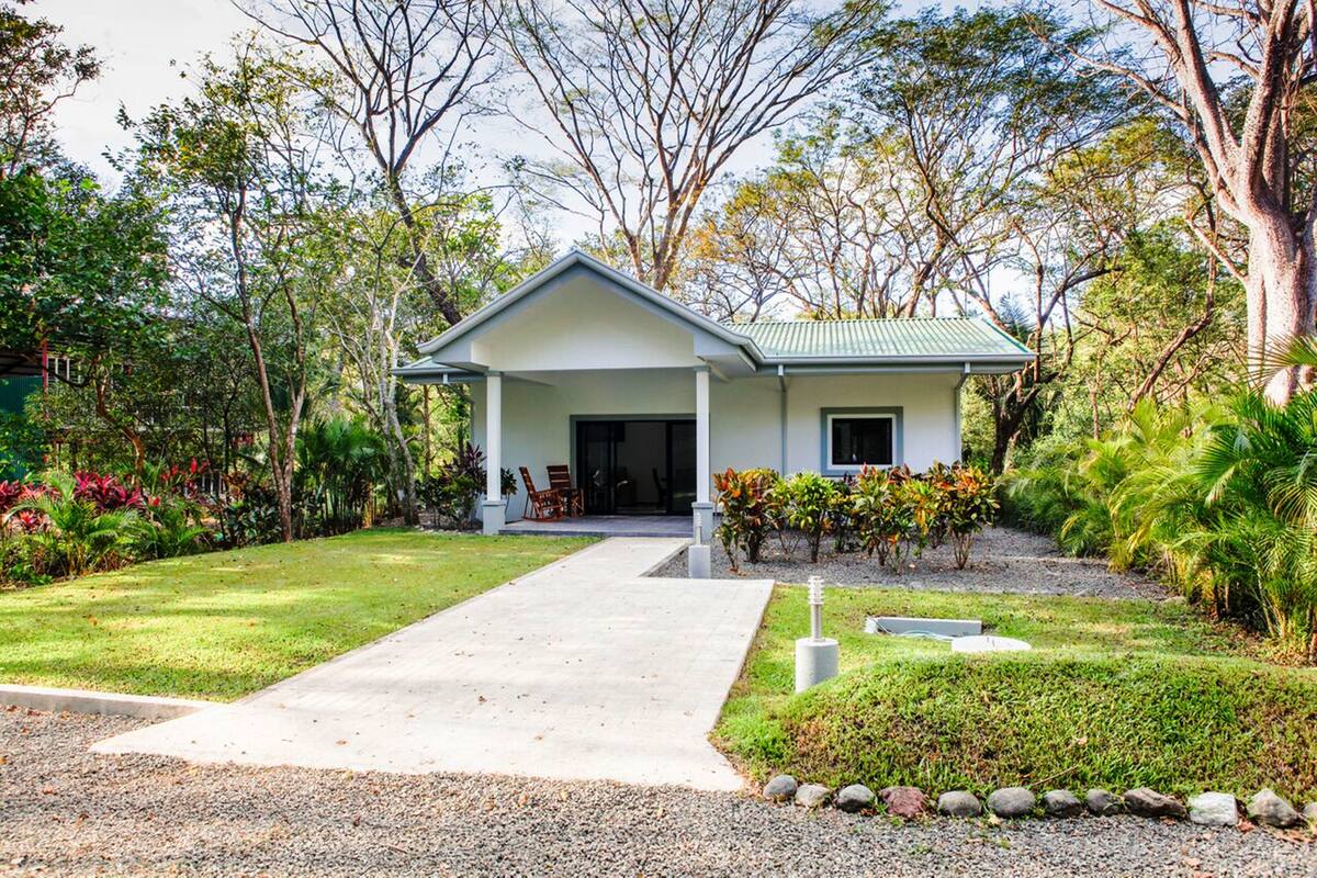 A modern house with a front porch, surrounded by lush greenery and a gravel driveway.